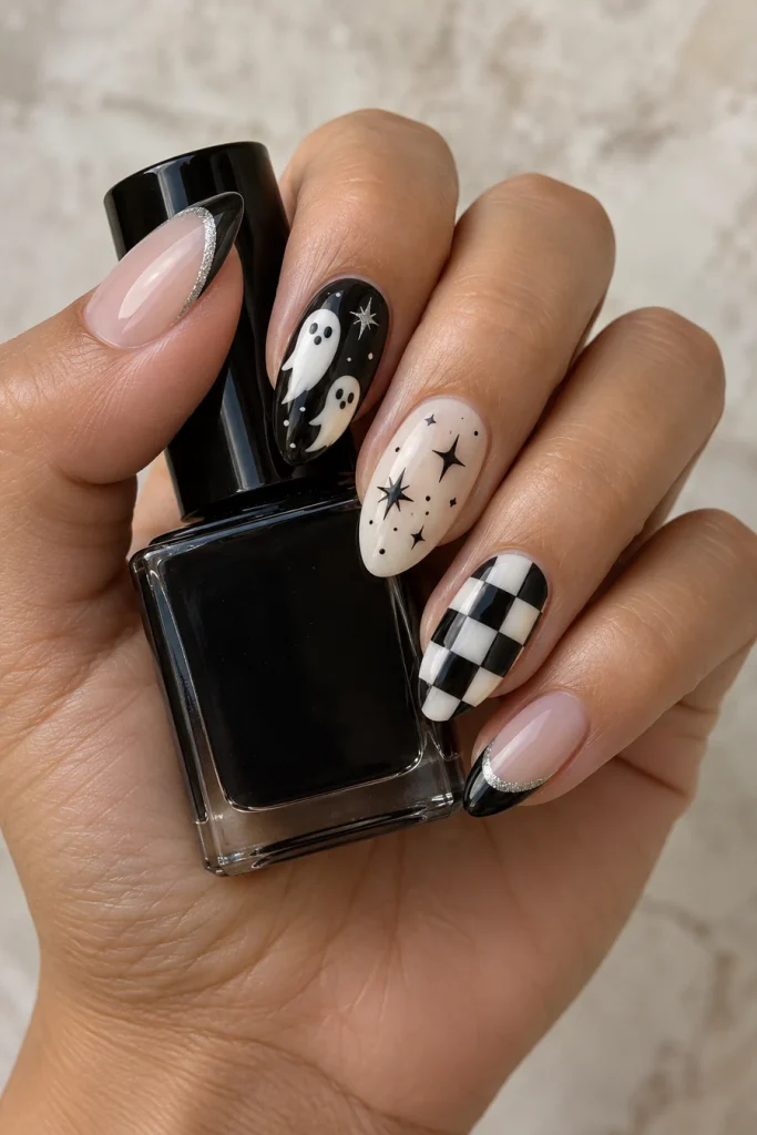 A hand holding a black bottle with almond nails featuring white ghosts, black stars on ivory, black-and-white checks and silver-edged black tips.