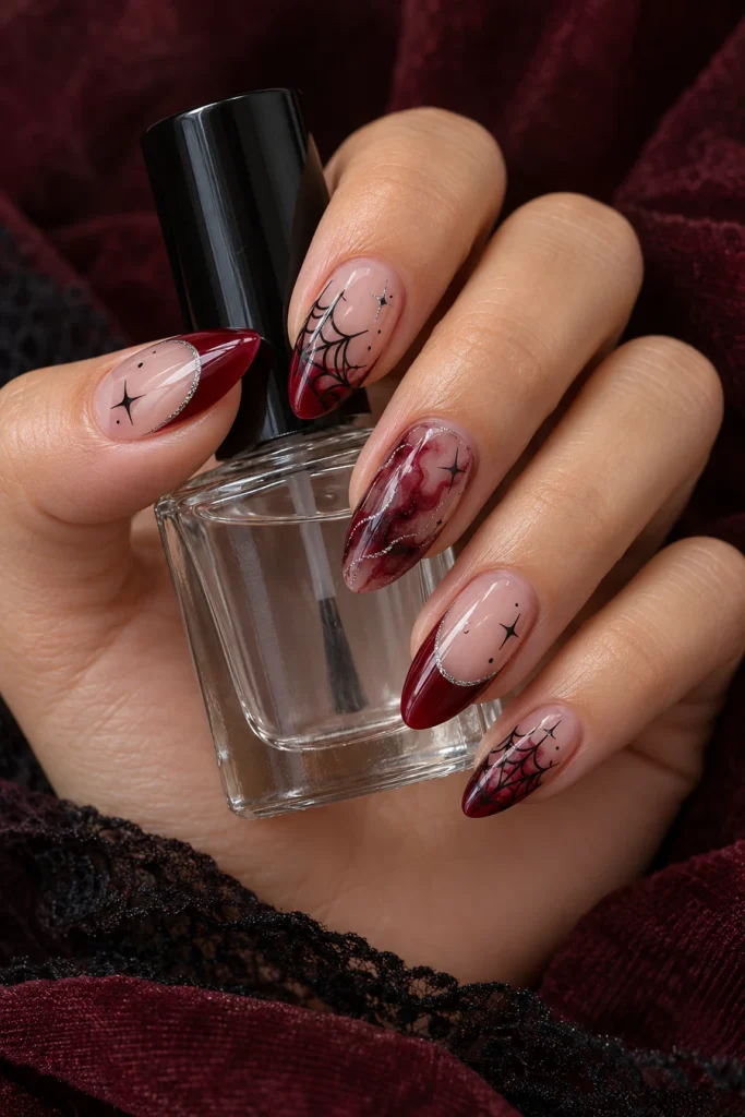 A hand holding a clear bottle with almond nails featuring burgundy French tips, black webs, smoky red marbling, small black stars and silver edges.